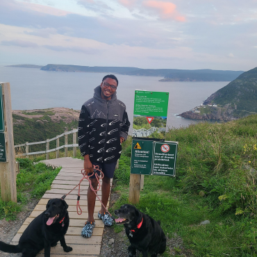 A young man stands on top of a hill with two large black dogs and the ocean behind him